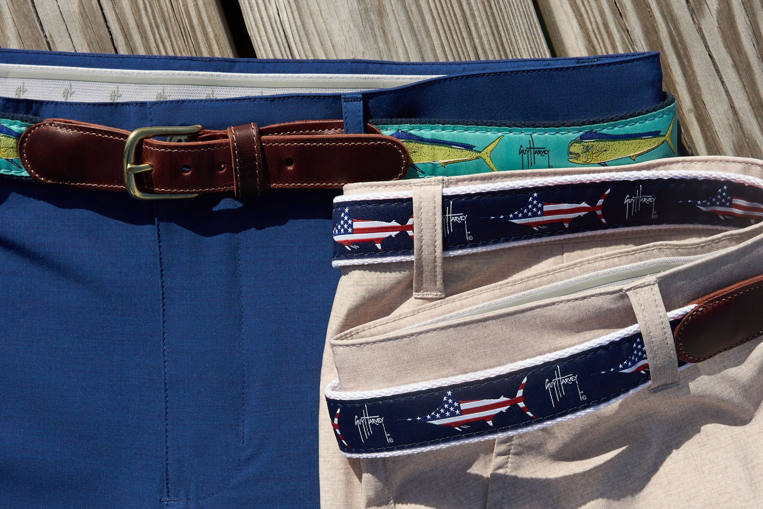 Two pairs of performance shorts with decorative leather tab belts featuring fish and American flag Guy Harvey designs.