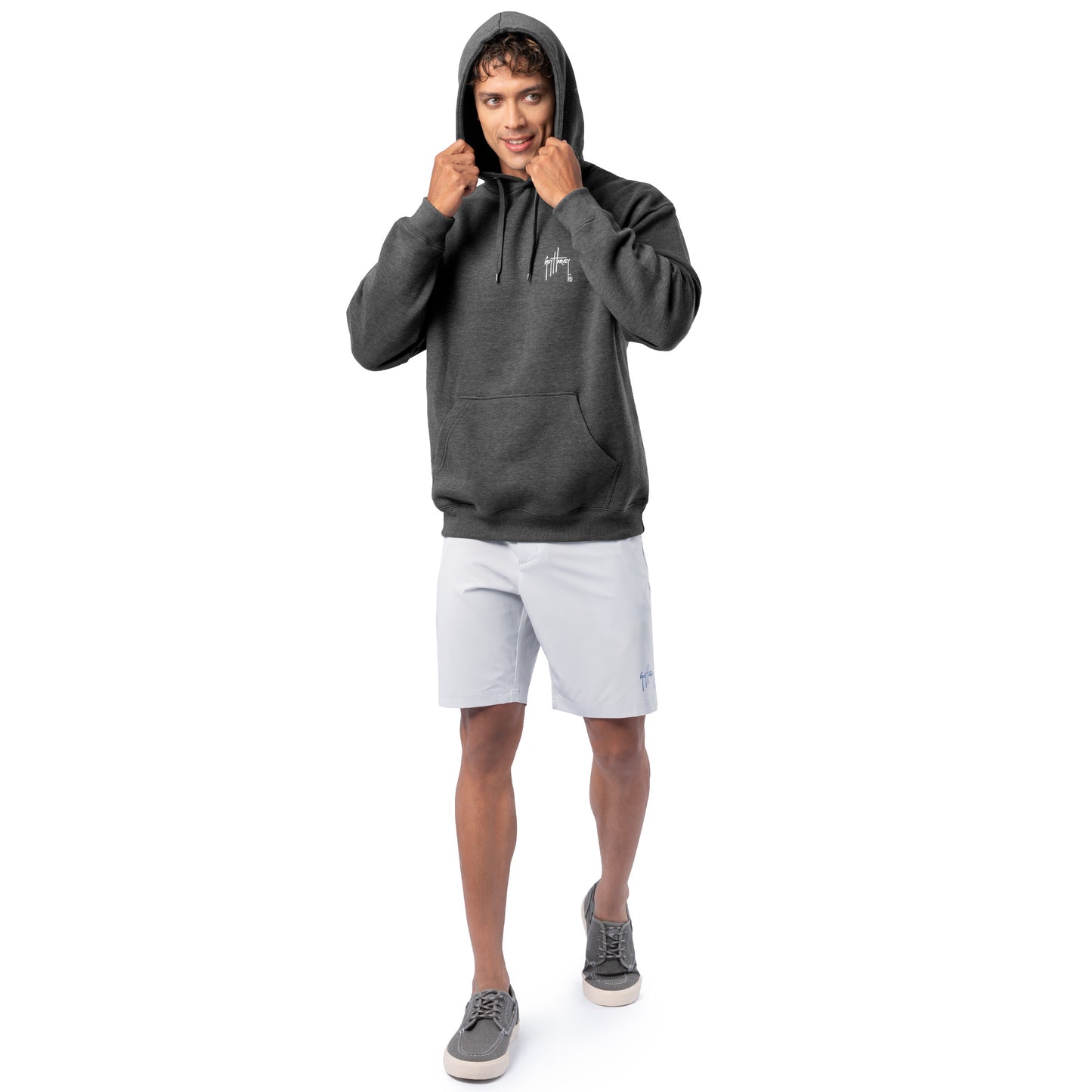 Full-body view of a man wearing a dark gray Guy Harvey fishing fleece hoodie with hood up and light shorts.