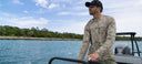 A man driving a fishing boat while wearing a camo Guy Harvey Marlin Air Sun Protection Top.
