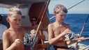 Two boys on a boat holding fishing reels with a clear blue sky and ocean background.