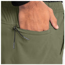 Close-up of olive green pants with a side pocket and a hand reaching into the pocket, showing a black drawstring with white 'Guy Harvey' text.