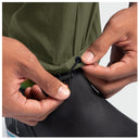Close-up of a person's hands fastening a black elastic compression cuff around the hem of an olive green technical fishing pant, paired with black deck boots.