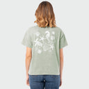 A woman viewed from behind wearing a light sage green t-shirt with a white seashell and starfish graphic on the back, paired with blue jeans, against a light gray background.