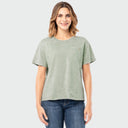 A woman modeling a light sage green short-sleeve t-shirt with a subtle mineral wash texture and a chest pocket, paired with blue jeans against a light gray background.