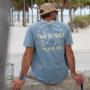 A man sits on a concrete ledge at a beach, viewed from behind, wearing a washed blue t-shirt with a large yellow and white graphic that reads 'TRAP TO TABLE' and 'THE BLUE CRAB'. A matching insulated tumbler with fish illustrations sits beside him.