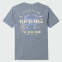 A heathered slate blue t-shirt showing the back graphic with a blue crab illustration and cream-colored text reading 'TRAP TO TABLE' and 'THE BLUE CRAB'.