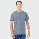 A man wearing a slate blue-gray short-sleeve pocket t-shirt with a small Guy Harvey logo on the chest pocket, paired with blue jeans, against a light gray background.