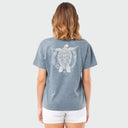 A woman viewed from behind wearing a faded blue-gray short-sleeve t-shirt with a large white sea turtle graphic on the back, paired with white shorts, against a light gray studio background.