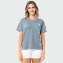 A woman modeling a short-sleeve crewneck t-shirt in a washed slate blue color, featuring a small chest pocket with a white logo tag on the left side. She is also wearing white shorts, set against a solid light gray background.