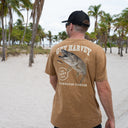 A man standing on a sandy beach with palm trees, viewed from behind, wearing a brown graphic t-shirt with a large fish design and the text 'GUY HARVEY' and 'THE FIFTY CLUB'.