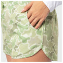 Close-up of a woman wearing light green and white Mallard Camo shorts, with a hand reaching into a side pocket. A small 'Guy Harvey' logo is visible on the front of the shorts.