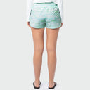 A woman shown from the back wearing light mint green and white patterned athletic shorts, a white long-sleeve top, and black flat sandals against a light gray studio background.