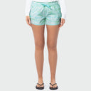 A women from the waist down wearing light green and blue tie-dye shorts, a white long-sleeve shirt, and black flip-flops against a light gray background.