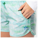 Close-up of a woman wearing mint green and white tie-dye swim shorts with a side pocket, shown from the waist down with a hand resting in the pocket. A blue script logo reading 'Guy Harvey' is visible on the lower left leg.