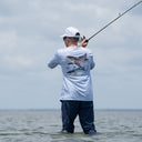 A man stands in shallow water wearing a light blue long-sleeve fishing shirt with a large fish graphic on the back, dark pants, and a white cap, holding a fishing rod under his arm.