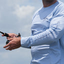A close-up of a man outdoors wearing a light blue long-sleeve performance shirt with black printed graphics on the chest and forearm, holding a fishing rod.