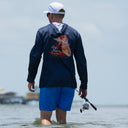 A man standing in shallow water on a beach, viewed from behind, wearing a navy blue long-sleeve fishing hoodie with a large redfish graphic and the text 'Guy Heavy' on the back, blue shorts, and a white cap.