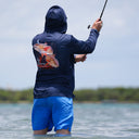 A man standing in shallow water wearing a navy blue long-sleeve fishing shirt with a large fish graphic on the back, paired with bright blue shorts, holding a fishing rod.