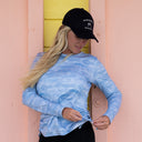 A woman wearing a light blue tie-dye long-sleeve sun protection top and a dark navy baseball cap with white embroidered text, standing against a pink and yellow wooden wall.