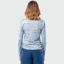 A woman viewed from behind wearing a light blue and white marbled long-sleeve sun protection top and dark blue pants against a light gray background.
