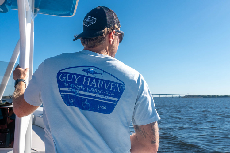 Man on a boat wearing a Guy Harvey shirt with a clear blue sky and water background.