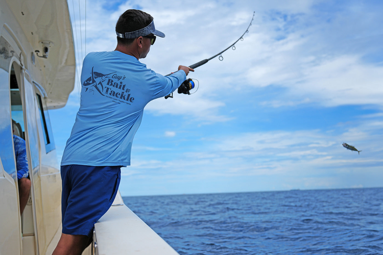 Man fishing on a boat wearing a Guy Harvey Eco Cast sun protection fishing shirt with a clear blue sky and ocean background