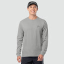 A man wearing a light gray long-sleeve crewneck shirt with a small black script logo on the left chest, paired with a dark gray baseball cap and dark pants, against a solid light gray background.