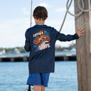 A boy stands on a dock by the water wearing a navy long-sleeve fishing shirt with a large back graphic that reads 'Catch a Wave' and blue shorts, holding a fishing rod and touching a wooden post with a rope.