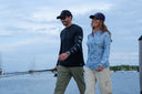A man and a woman walking outdoors by the water wearing matching long-sleeve performance fishing shirts. The man wears a black shirt with a subtle white fish pattern on the sleeve, and the woman wears a light blue patterned shirt.