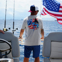 Man on a boat wearing a gray t-shirt with a marlin design, blue shorts, and a cap with an American flag, looking out over water.