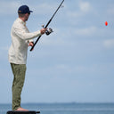A man wearing an Angler Edge performance fishing pant in color green and a button down Eco Angler fishing shirt in color almond milk from Guy Harvey. He is standing on a boat about to cast.