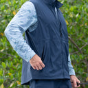 A man wearing a navy blue zip-up fishing vest over a light blue camouflage long-sleeve shirt, standing outdoors in front of green foliage.