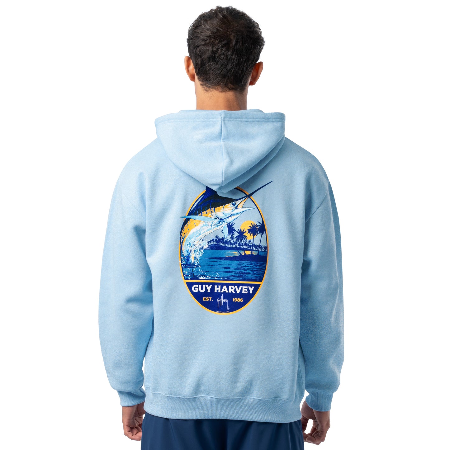 Rear angled view of a man wearing a light blue Guy Harvey fishing fleece hoodie featuring marlin and tropical artwork