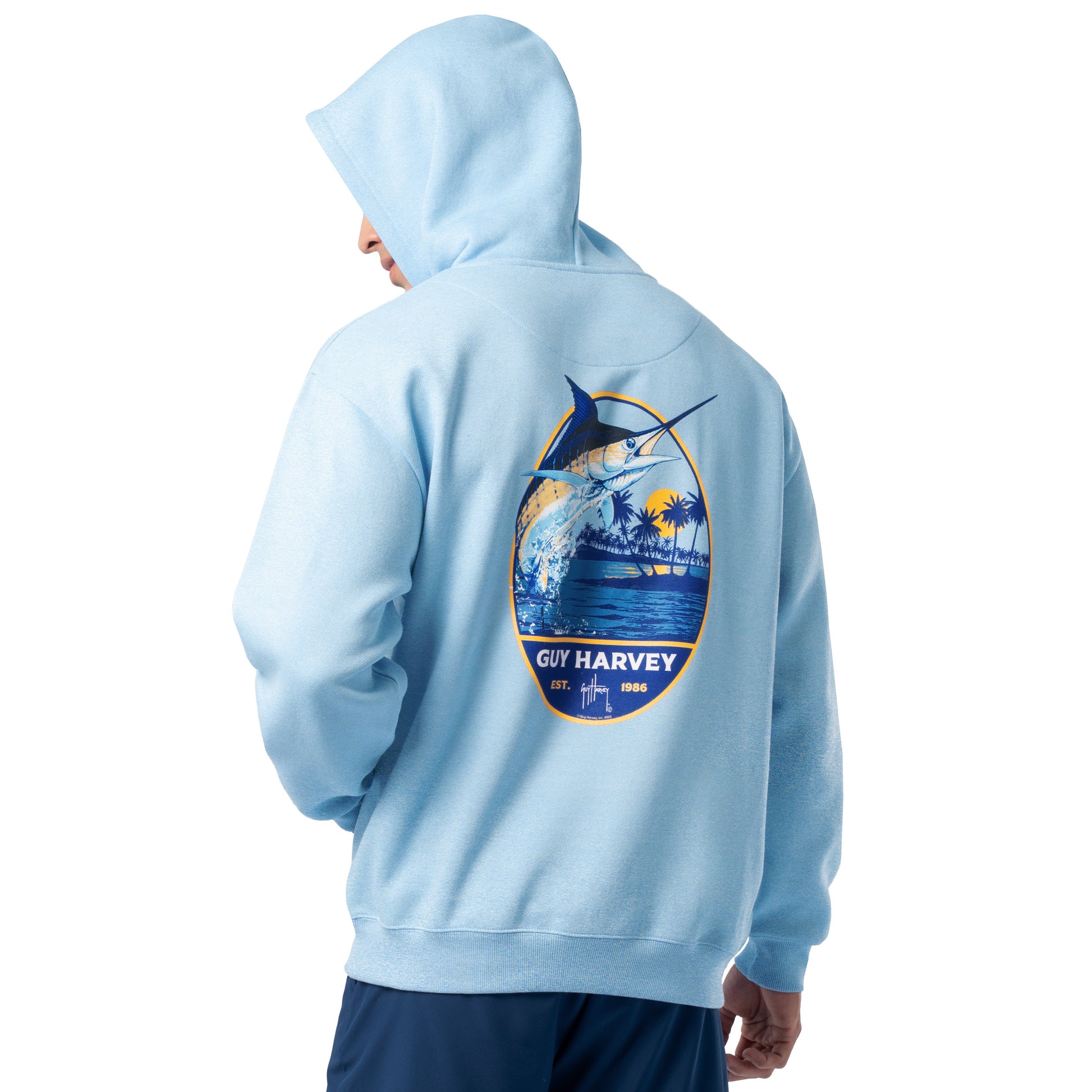 Back view of a man wearing a light blue Guy Harvey fishing fleece hoodie featuring a colorful marlin jumping artwork with palm trees and ocean scene.