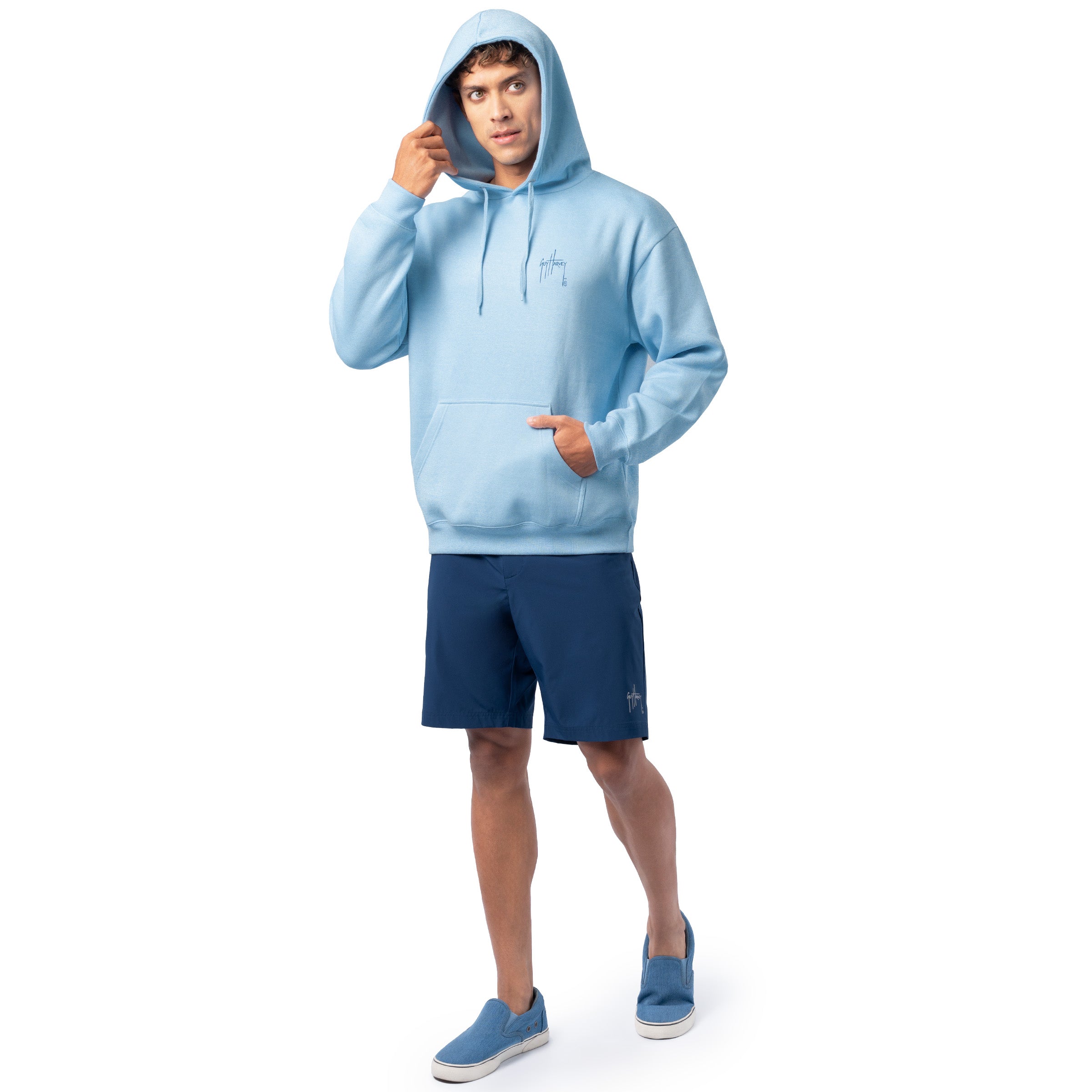 Full-body view of a man wearing a light blue Guy Harvey fishing fleece hoodie with hood up and navy shorts.