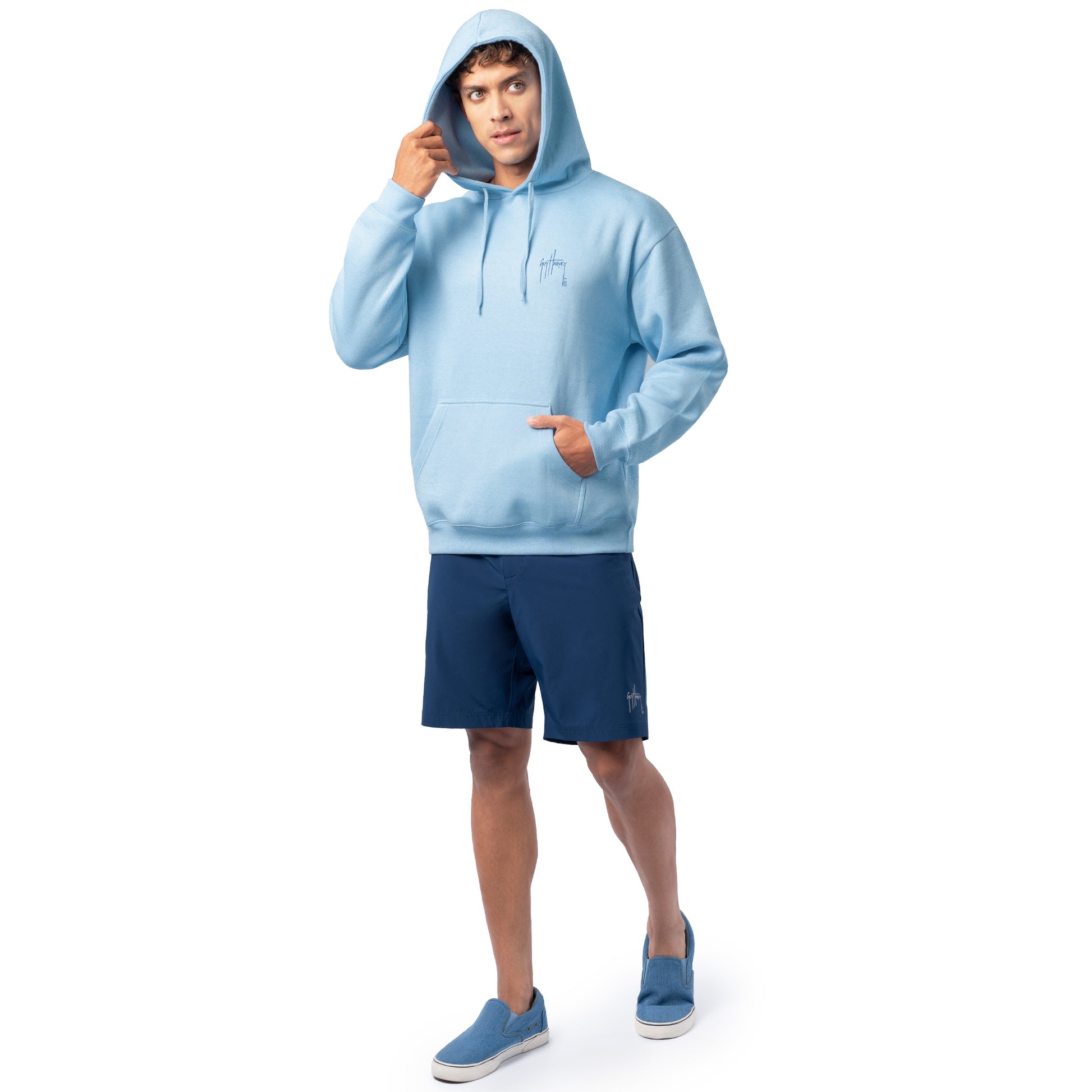 Full-body view of a man wearing a light blue Guy Harvey fishing fleece hoodie with hood up and navy shorts.