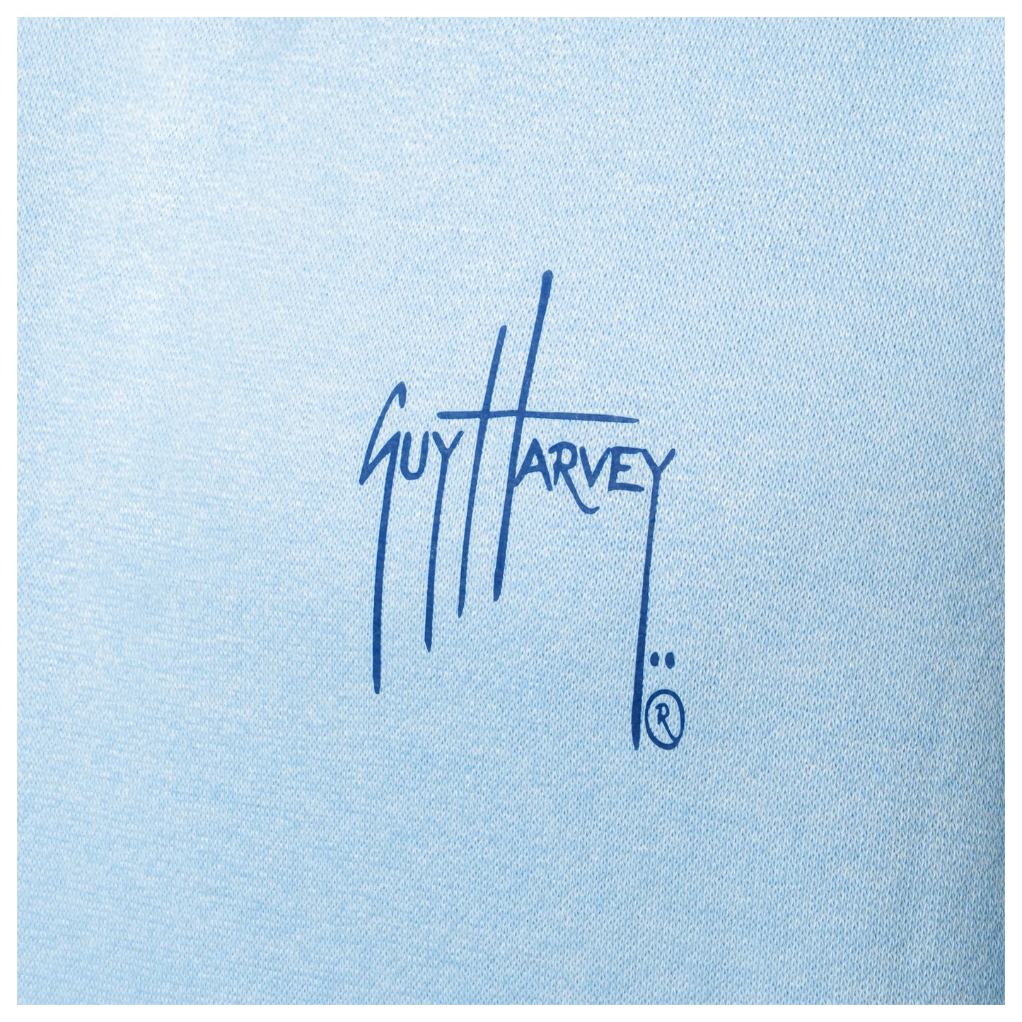 Macro close-up of the blue Guy Harvey signature logo on the fabric of a light blue fishing fleece hoodie.