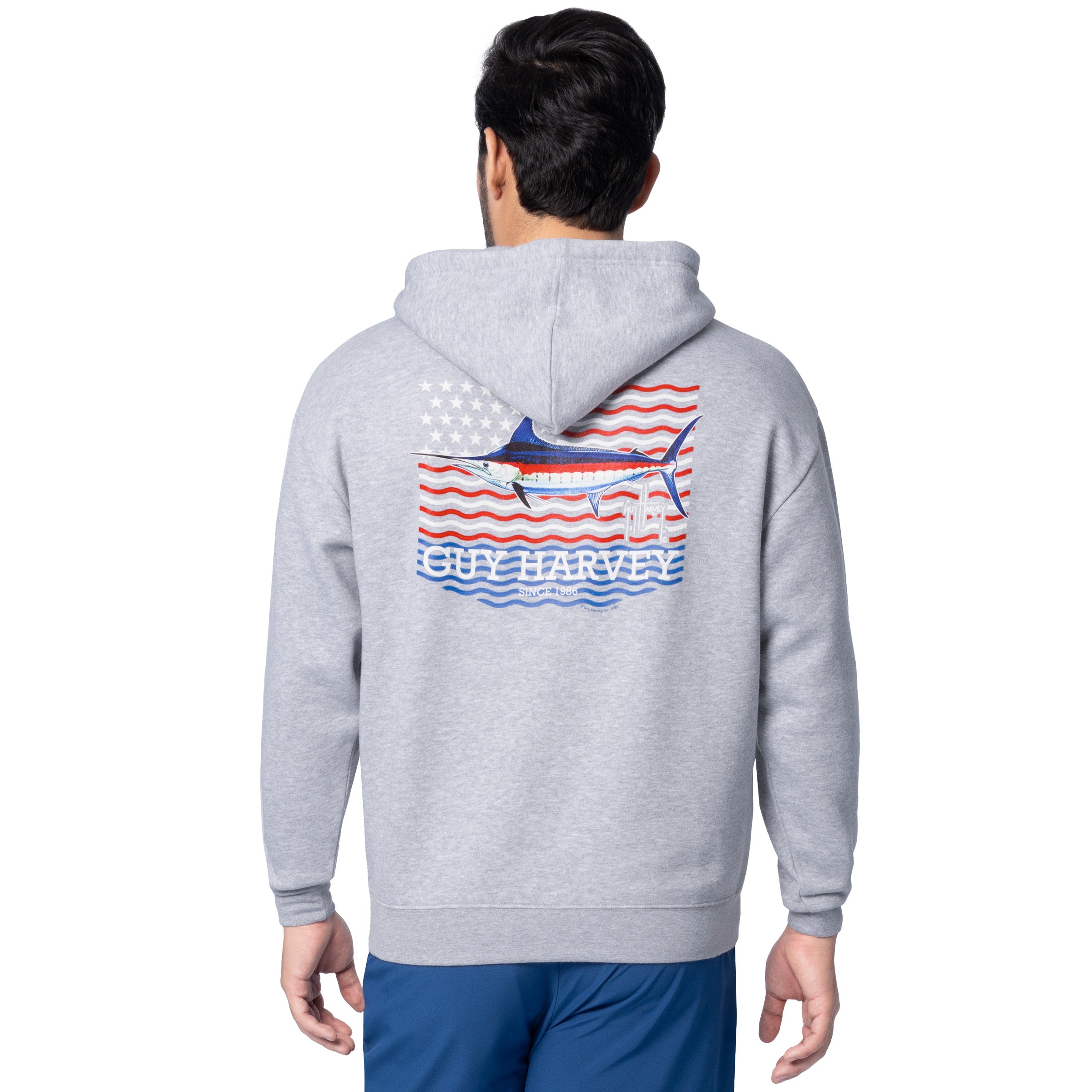 Rear angled view of a man wearing a light gray Guy Harvey fishing fleece hoodie featuring marlin and USA flag artwork.