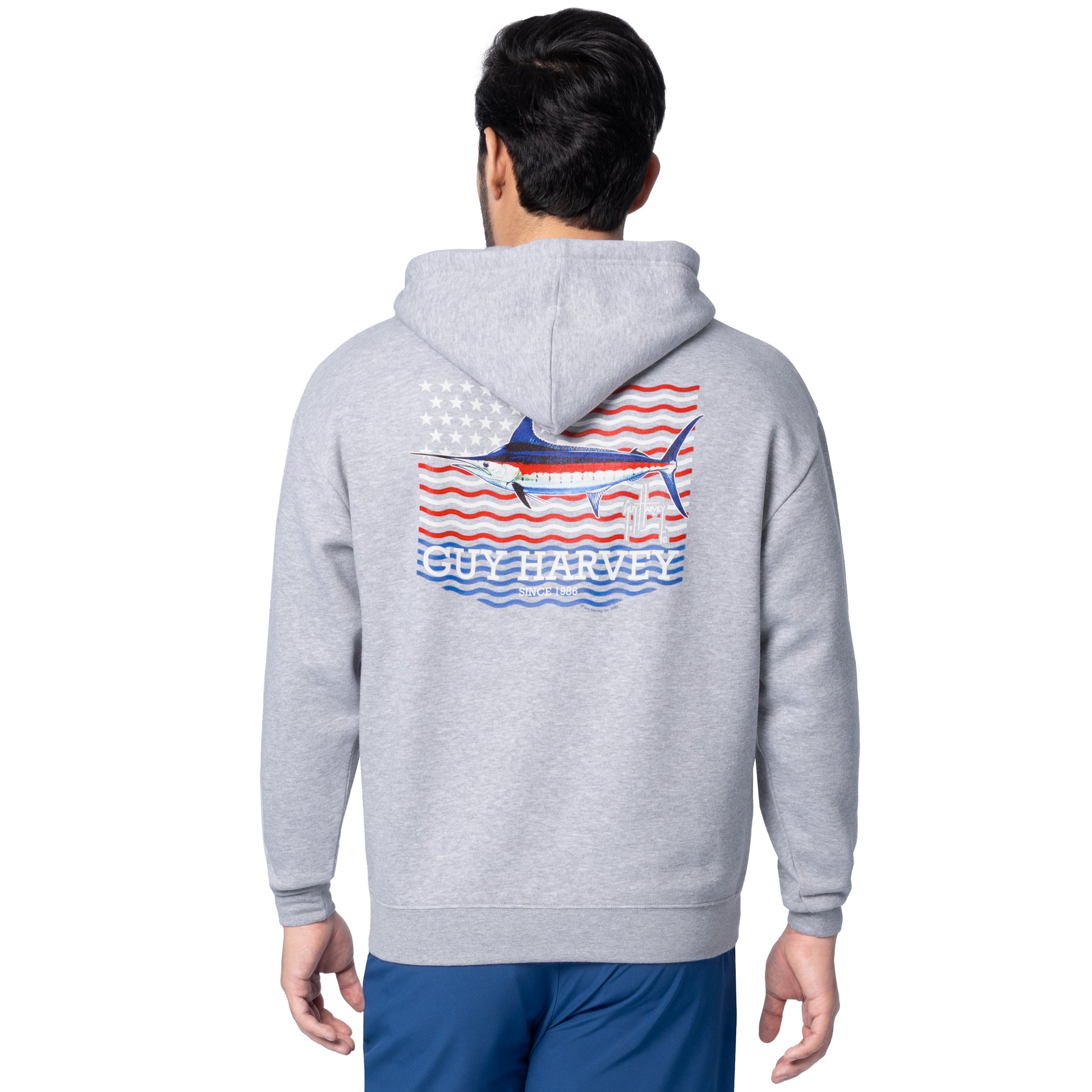 Rear angled view of a man wearing a light gray Guy Harvey fishing fleece hoodie featuring marlin and USA flag artwork.