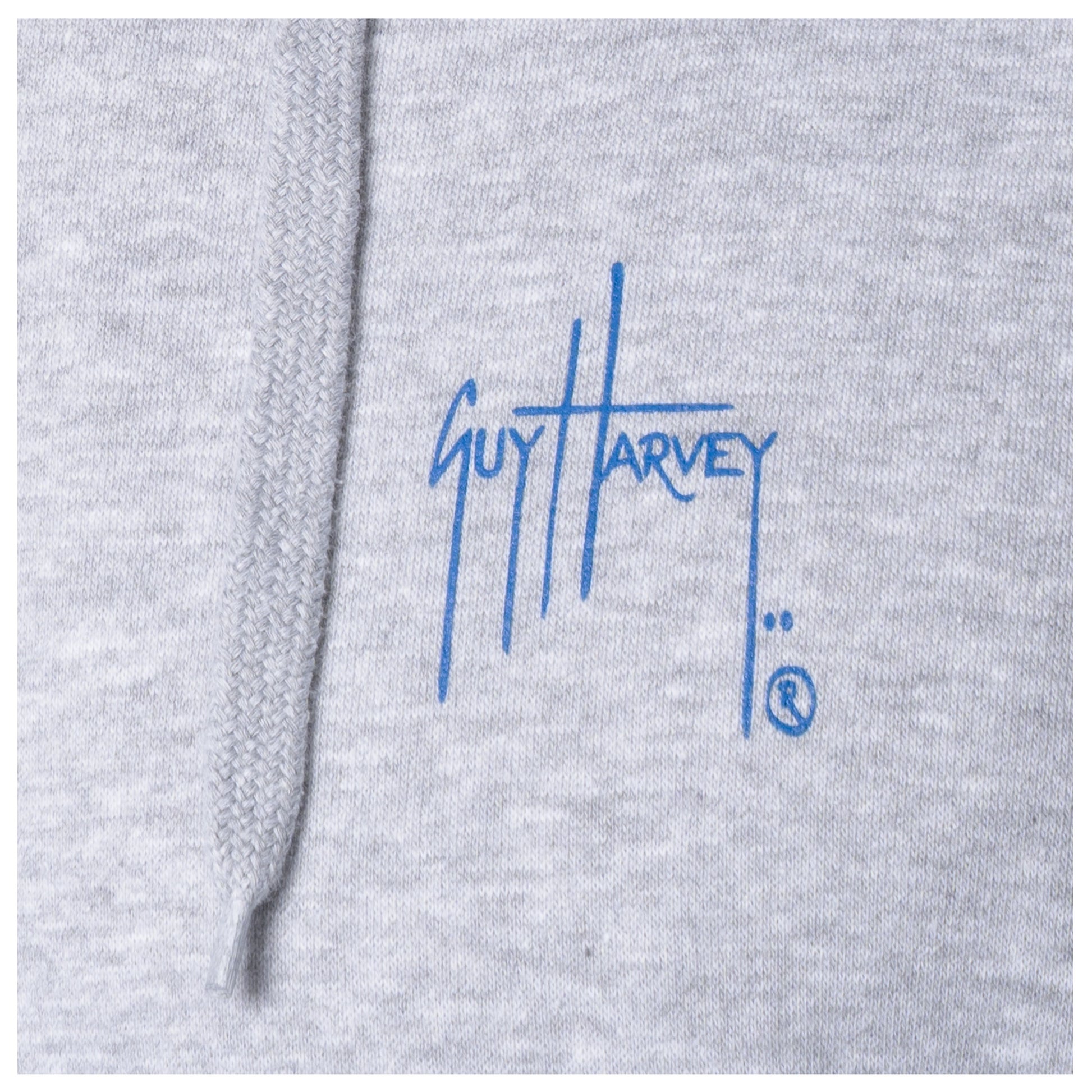 Macro close-up of the printed Guy Harvey signature logo on the fabric of a light gray fishing fleece hoodie.