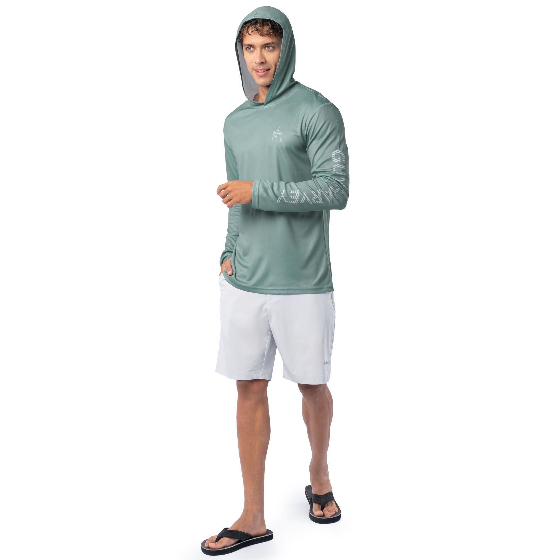 Lifestyle image of model wearing seafoam green Guy Harvey Men’s Mahi Gear Sun Protection Hoodie with hood up and white shorts.