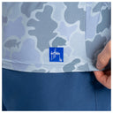 Close-up of a person wearing a light blue and gray camouflage patterned top with a blue square patch featuring the brand name 'Guy Harvey' on the bottom left, paired with solid blue bottoms.