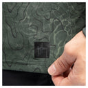 Close-up of a person wearing a dark green top with an topographic wavy line pattern and a black brand patch reading 'GUY HARVEY' on the bottom left.