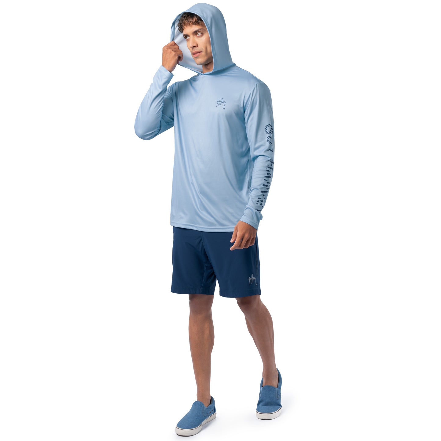 Lifestyle image of model wearing light blue Guy Harvey Men’s Sail Stripes Sun Protection Hoodie with navy shorts.