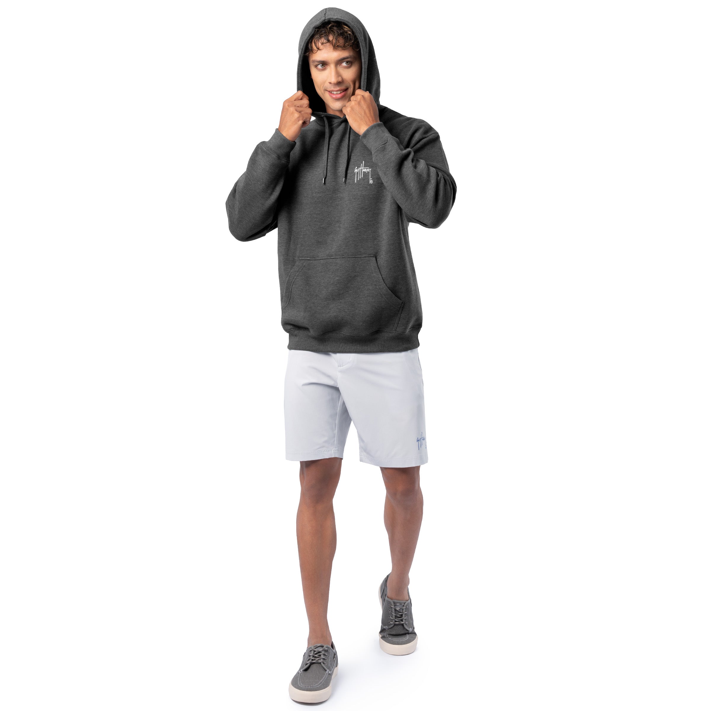 Full-body view of a man wearing a dark gray Guy Harvey fishing fleece hoodie with hood up and light shorts.