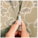 Close-up of a person's hand pulling a silver zipper tab on a camouflage zip-up jacket. The zipper pull is engraved with the brand name 'GUY HARVEY'.