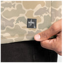 Close-up of a camouflage-patterned garment with a visible black Guy Harvey logo patch, held by a hand against a dark fabric background.