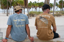 Two men sitting on a concrete ledge at a beach, seen from behind. The man on the left wears a light blue t-shirt with a large fish graphic and 'TRAP TO TABLE' text, while the man on the right wears a brown t-shirt with a large bass graphic and 'GUY HARVEY' text.