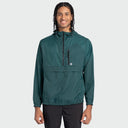 A man wearing a dark teal green hooded windbreaker jacket with a half-zip front, a horizontal chest zip pocket, and elastic cuffs, paired with black pants, against a light gray background.