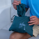 A person in a blue shirt holds a dark teal drawstring pouch with a white script logo that reads 'Guy Harvey'. The pouch is shown in a close-up shot while the person sits on a white cushioned surface.